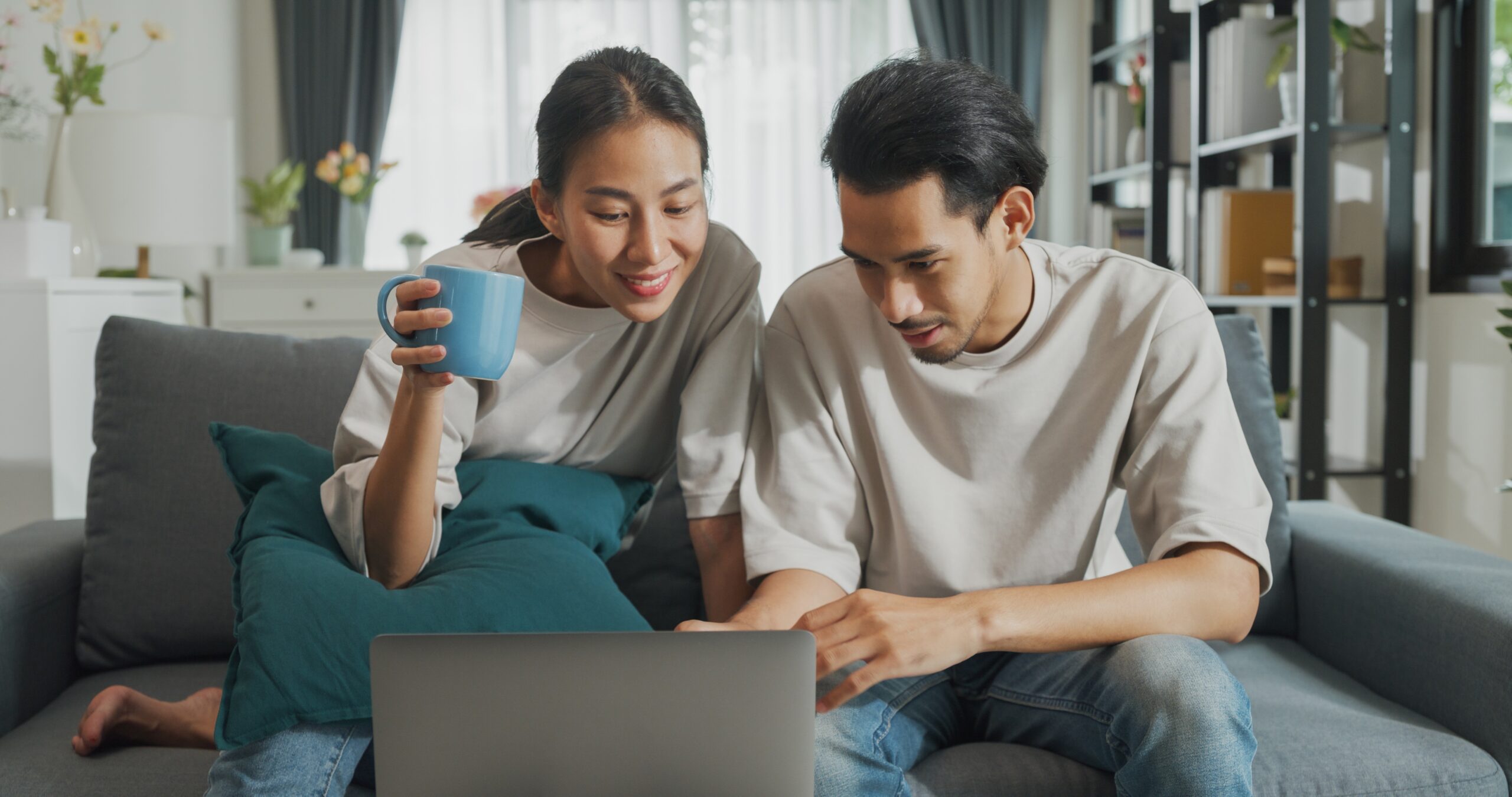 Happy young Asian couple sit on couch looking at laptop