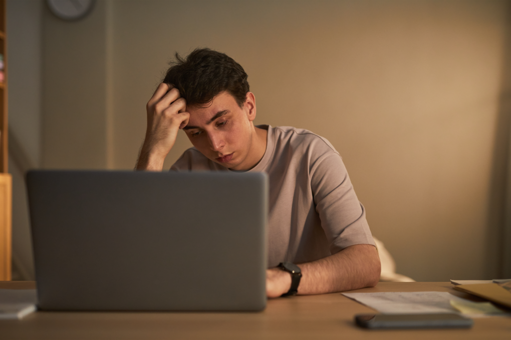 Young man sitting at desk holding head in hand, while looking at laptop screen